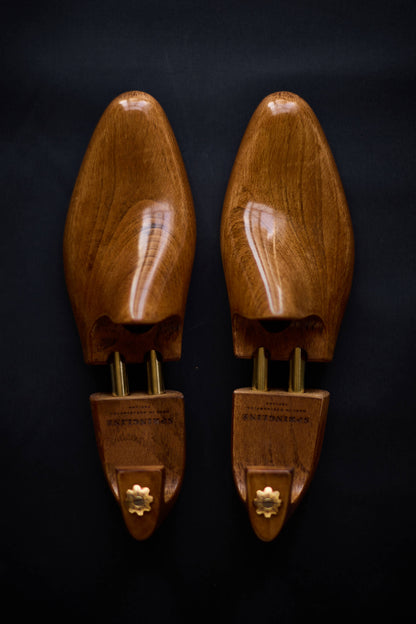 A pair of brown wooden shoe trees with metal hinges displayed against a dark background.